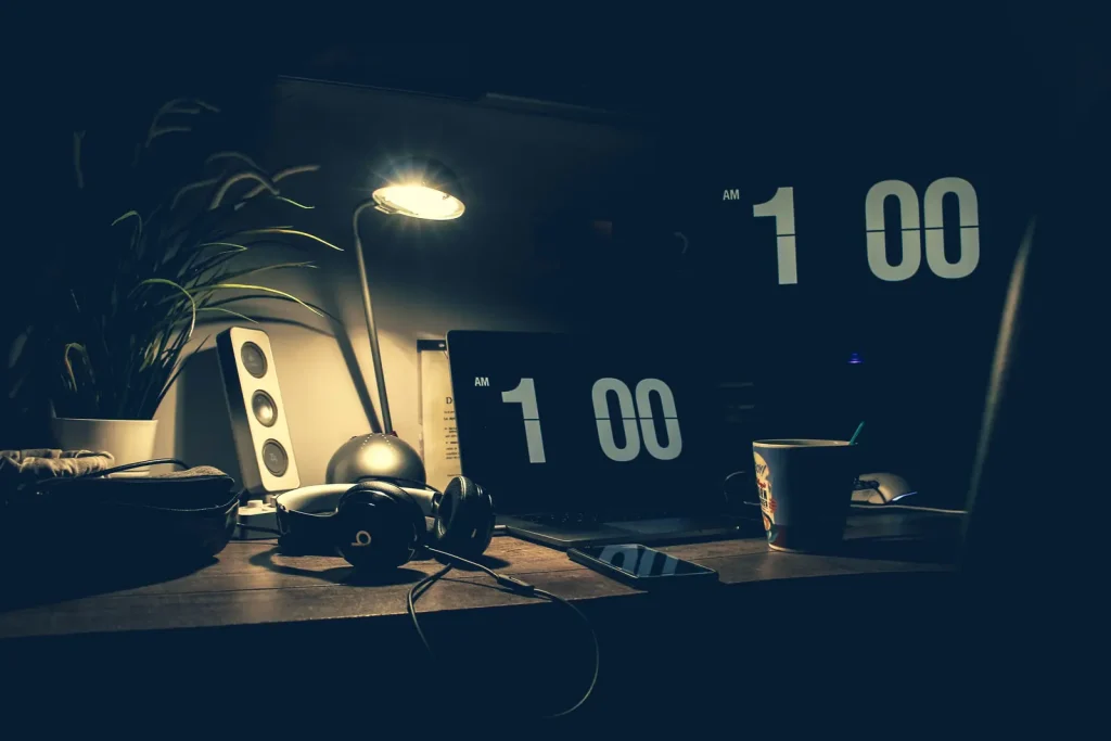 Birou slab luminat noaptea, cu un laptop, o lampă și un ceas